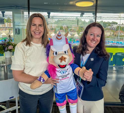 Two women standing in a sports venue. They are both smiling towards the camera, and are holding Finnie, the soft toy mascot of the Glasgow 2026 Commonwealth Games