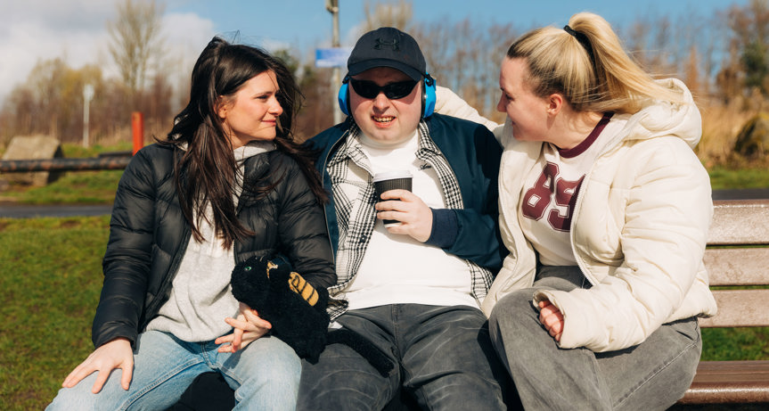 Three people sitting on a park bench in a country park. On the left is a young woman with long dark hair, wearing jeans and a black puffer jacket over a grey hoodie. In the middle is a young man wearing a black baseball cap, sunglasses and headphones. He is wearing black jeans, a black and white striped shirt over a white t-shirt, and a dark blue jacket. The young woman on the right has long blonde hair in a ponytail and is wearing dark grey jeans with a cream puffer jacket over a cream t-shirt