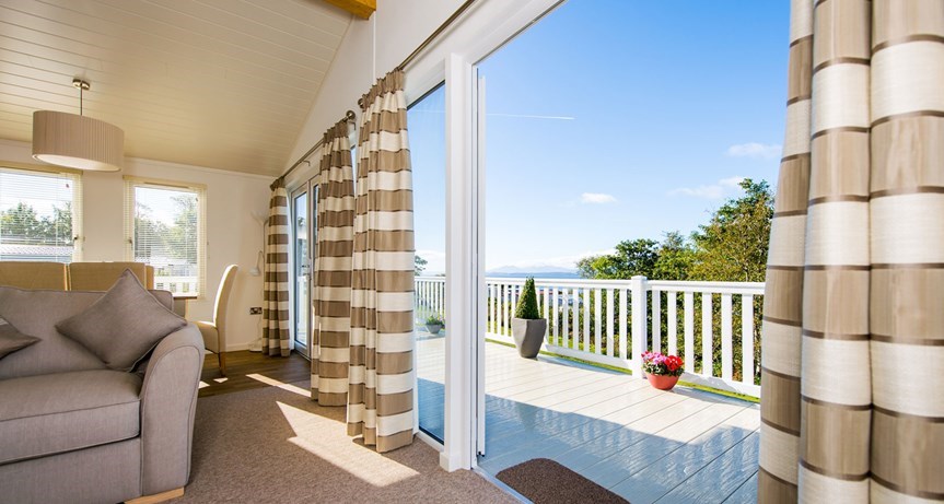 An interior shot of a lounge area in a holiday lodge. As well as seeing some soft furnishings, the patio doors are open there is a view out over a decking area to some trees. The sky is blue with very few clouds.