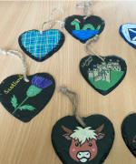Hand-painted slate hearts featuring Scottish-themed designs including tartan, the Loch Ness Monster, Edinburgh Castle and a Highland Cow