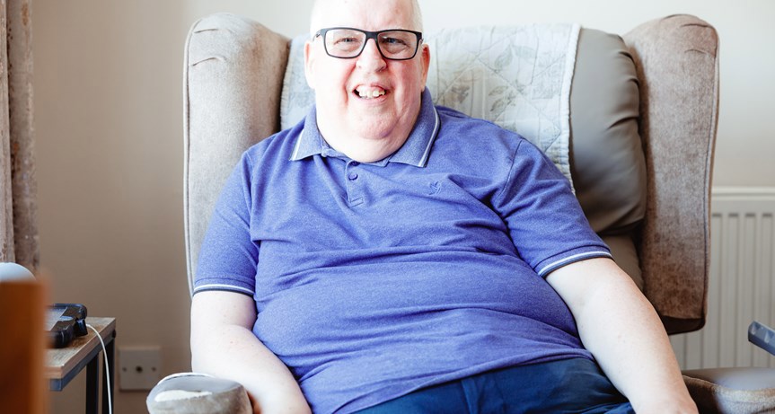 Man sitting on an armchair in his home. He is smiling towards the camera, wears glasses and a purple polo shirt.