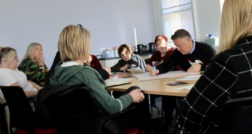 A group people sitting at a meeting table. They are in discussion and someone is writing on a flipchart.